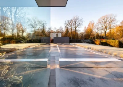 alice-et-paul-gallery-01 Allée en béton avec des reflets de verre, bordée de haies et d'arbres nus sous un ciel clair et ensoleillé.