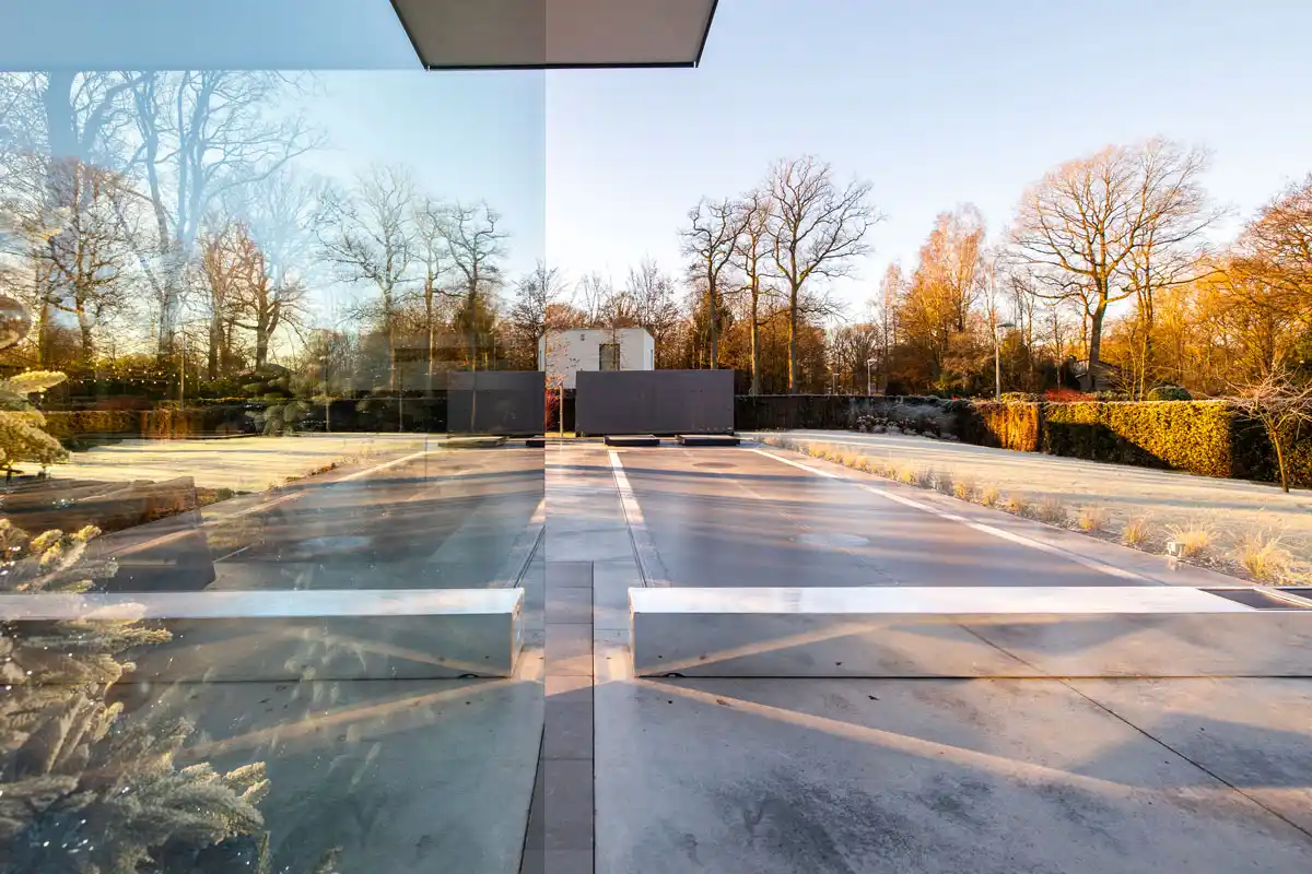 Allée en béton avec des reflets de verre, bordée de haies et d'arbres nus sous un ciel clair et ensoleillé.