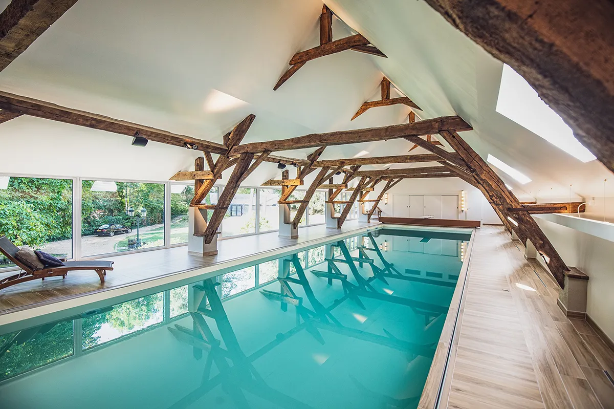 Piscine intérieure avec eau turquoise, entourée de poutres en bois apparentes sous un toit incliné, avec vue sur le jardin extérieur.
