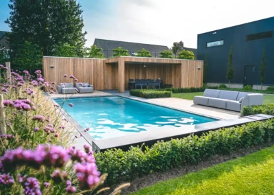 lete-chez-veerle-jan Piscine entourée de haies et de fleurs violettes, avec un salon de jardin en bois et des canapés gris sur la terrasse.