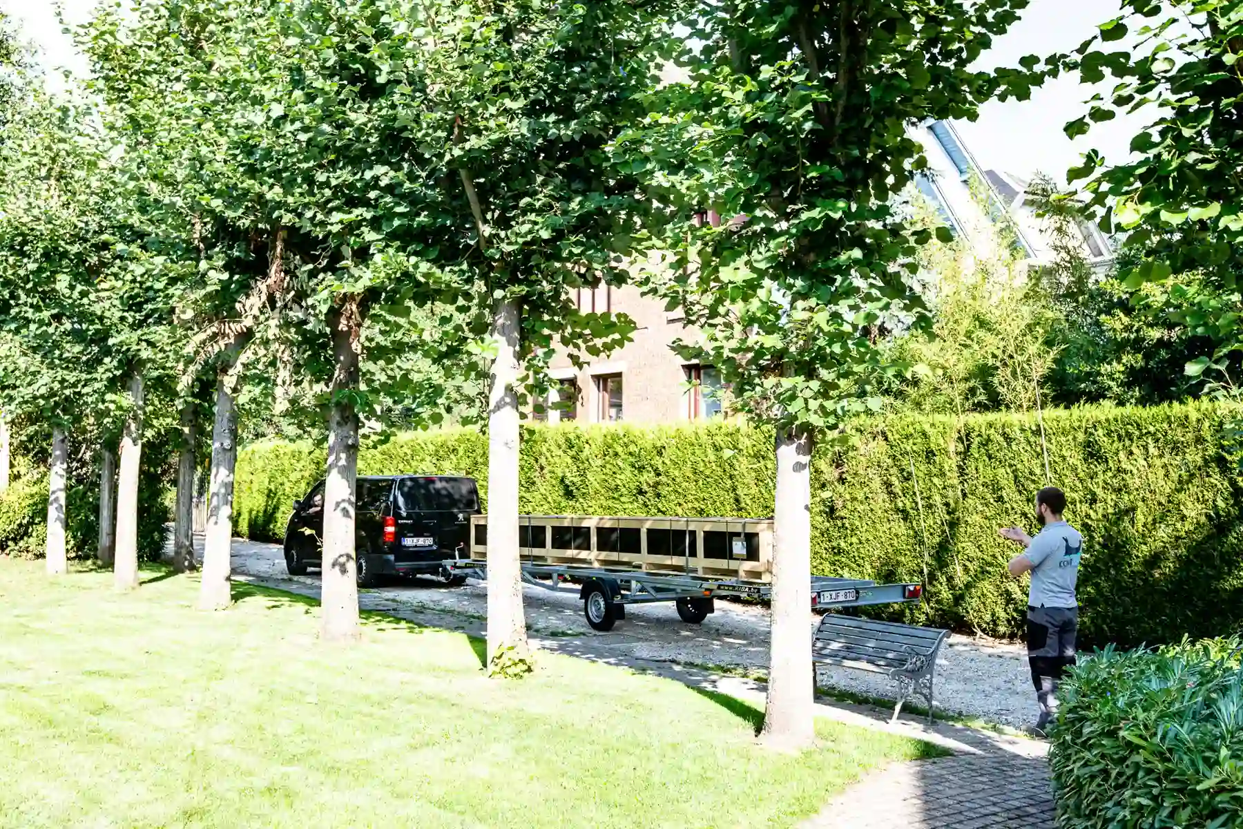 Un véhicule noir avec une remorque est stationné sur une allée bordée d'arbres, près d'une haie verte et d'un banc en métal.
