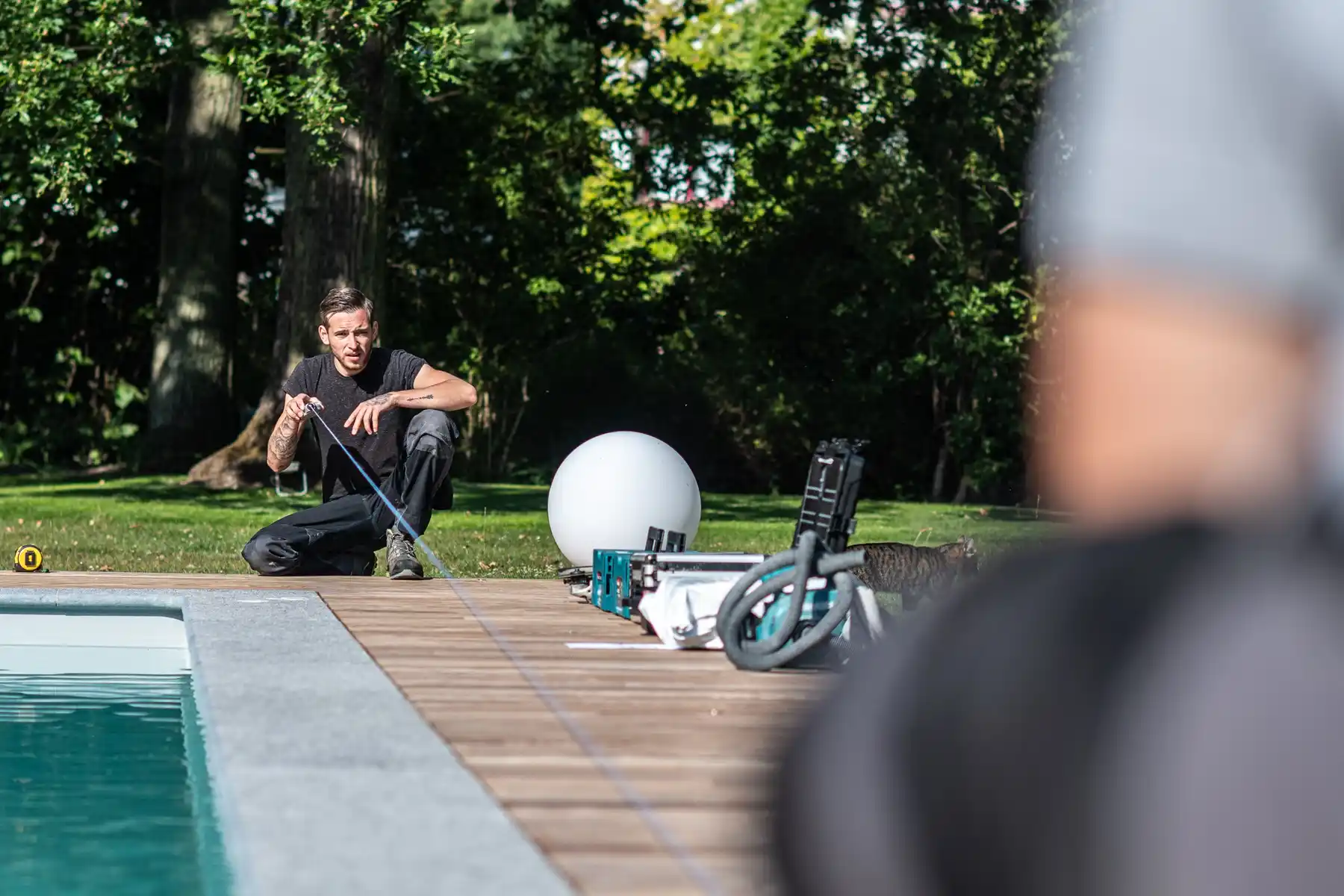 Un homme agenouillé mesure le bord d'une piscine avec un ruban à mesurer, entouré d'arbres et d'équipements divers.