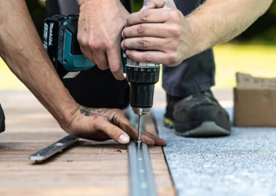 linstallation-chez-juliette-leon-3 Deux mains installent une vis avec une perceuse sans fil sur un rail métallique posé sur une surface en bois et tapis gris.