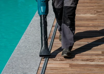 linstallation-chez-juliette-leon-6 Un homme en pantalon noir utilise un souffleur de feuilles bleu sur une terrasse en bois près d'une piscine turquoise.