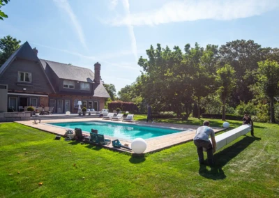 linstallation-chez-juliette-leon-9 Deux personnes installent une couverture de piscine blanche près d'une maison en briques avec un grand jardin verdoyant.