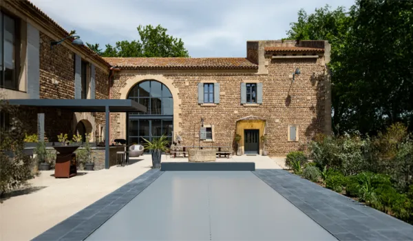 Cour intérieure avec piscine couverte, entourée de bâtiments en pierre, mobilier de jardin moderne et végétation luxuriante sous ciel nuageux.