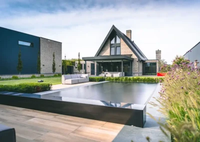 veerle-et-jan-besoins Maison moderne avec toit en pente entourée de jardin, piscine couverte au premier plan, et canapé extérieur gris clair sur la terrasse.