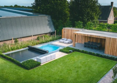 veerle-et-jan-coverseal Une piscine avec couverture grise est entourée de mobilier de jardin et d'une terrasse en bois, avec pelouse verte et haies soignées.