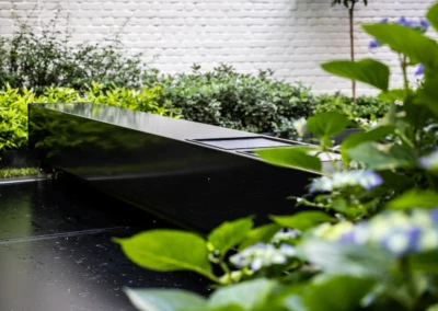 Une couverture de piscine noire entourée de plantes vertes et de fleurs violettes, avec un mur en briques blanches en arrière-plan.