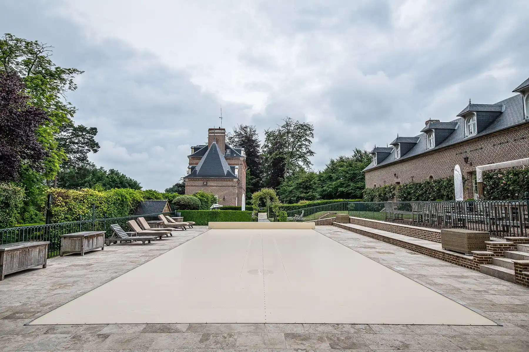 Piscine couverte de beige entourée de chaises longues, située devant un bâtiment en briques avec un ciel nuageux.