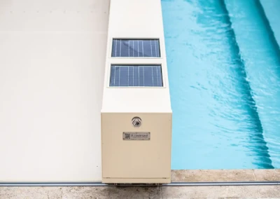 Boîtier blanc avec panneaux solaires à côté d'une piscine bleue, sur un sol en pierre beige.