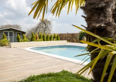 Chez-Charlotte-et-Pierre-1 Une piscine entourée de dalles en bois, avec un palmier au premier plan et un cabanon gris en arrière-plan.