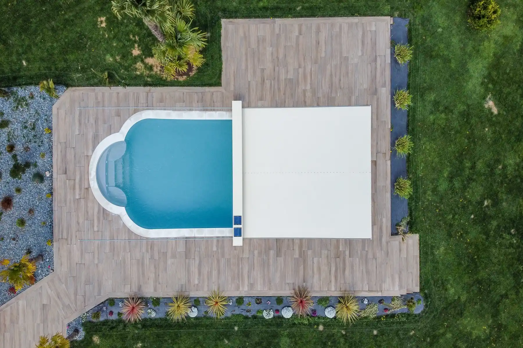 Vue aérienne d'une piscine rectangulaire avec couverture blanche, entourée de terrasse en bois et de pelouse verte.