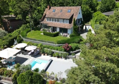Chez-Elena-et-Nathan-1 Maison avec toit en tuiles entourée de verdure, piscine et terrasse avec parasols blancs au premier plan.