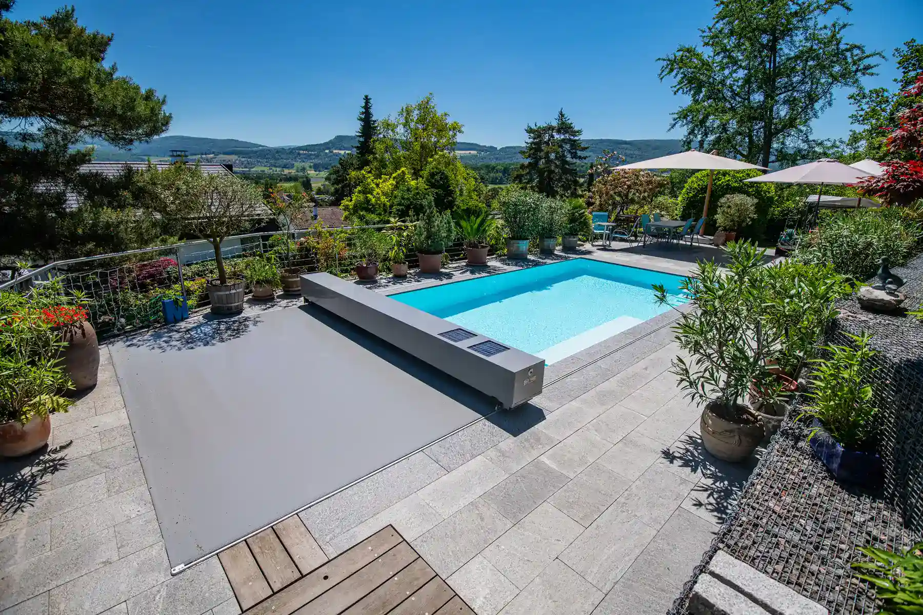 Piscine rectangulaire entourée de plantes en pot, avec une terrasse en dalles grises et parasols blancs sous un ciel bleu.