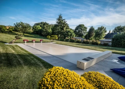 Chez-Gwendoline-et-François-1 Couverture de piscine beige entourée de pelouse verte, avec des pots de fleurs roses et des arbres en arrière-plan.