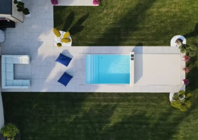 Chez-Gwendoline-et-François-2 Vue aérienne d'une piscine rectangulaire, entourée de dalles blanches, avec des coussins bleus et des arbustes décoratifs sur pelouse verte.