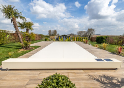 PALMIERS (11) Terrasse en bois avec une couverture de piscine blanche, entourée de palmiers et d'arbustes, menant à un abri de jardin sous un ciel partiellement nuageux.