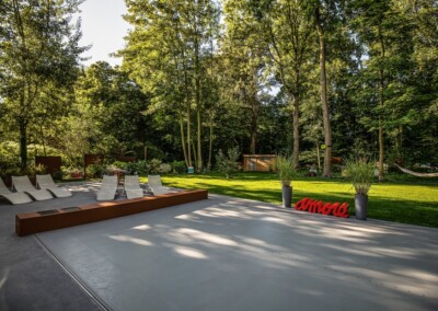 Des chaises longues blanches sont disposées sur une terrasse en bois, entourée de pelouse verte et d'arbres, avec des pots de fleurs noirs.
