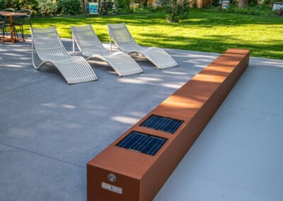 Trois chaises longues blanches sur une terrasse en béton, près d'un boîtier en métal rouillé avec panneaux solaires, entourées de verdure.