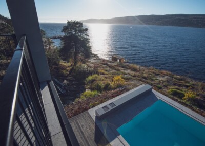 DROBAK - SUN (8) Vue d'un balcon surplombant une piscine bleue bordée de dalles grises, avec un paysage côtier et un arbre en arrière-plan.