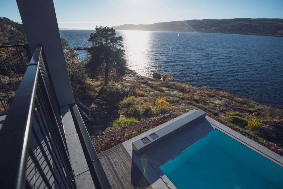 Vue d'un balcon surplombant une piscine bleue bordée de dalles grises, avec un paysage côtier et un arbre en arrière-plan.