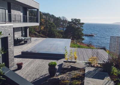 Terrasse en bois moderne avec piscine couverte, vue sur la mer et entourée de verdure et d'arbustes, sous un ciel bleu dégagé.