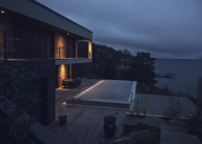 Maison moderne avec terrasse en bois éclairée, surplombant la mer, entourée de végétation et ciel nuageux.