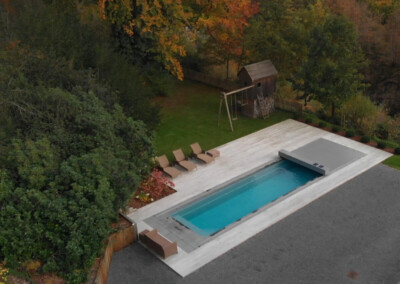Piscine rectangulaire entourée de chaises longues beige sur une terrasse en bois, avec un jardin et des arbres en arrière-plan.