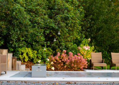 Des chaises en osier marron et un coffre gris sont disposés sur une terrasse en bois, entourés de buissons verts et de fleurs roses.