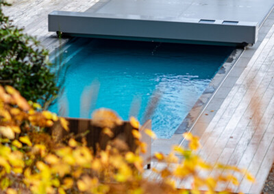 Une piscine bleue partiellement couverte par une structure grise, entourée de végétation automnale et d'une terrasse en bois clair.