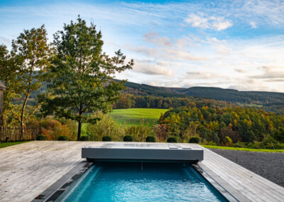 Piscine couverte sur terrasse en bois avec vue sur paysage vallonné et arbres verdoyants sous un ciel partiellement nuageux.