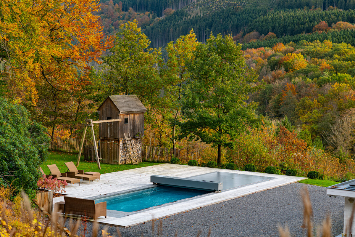 Une piscine rectangulaire avec couverture grise est entourée de chaises longues et d'un abri en bois, en automne.