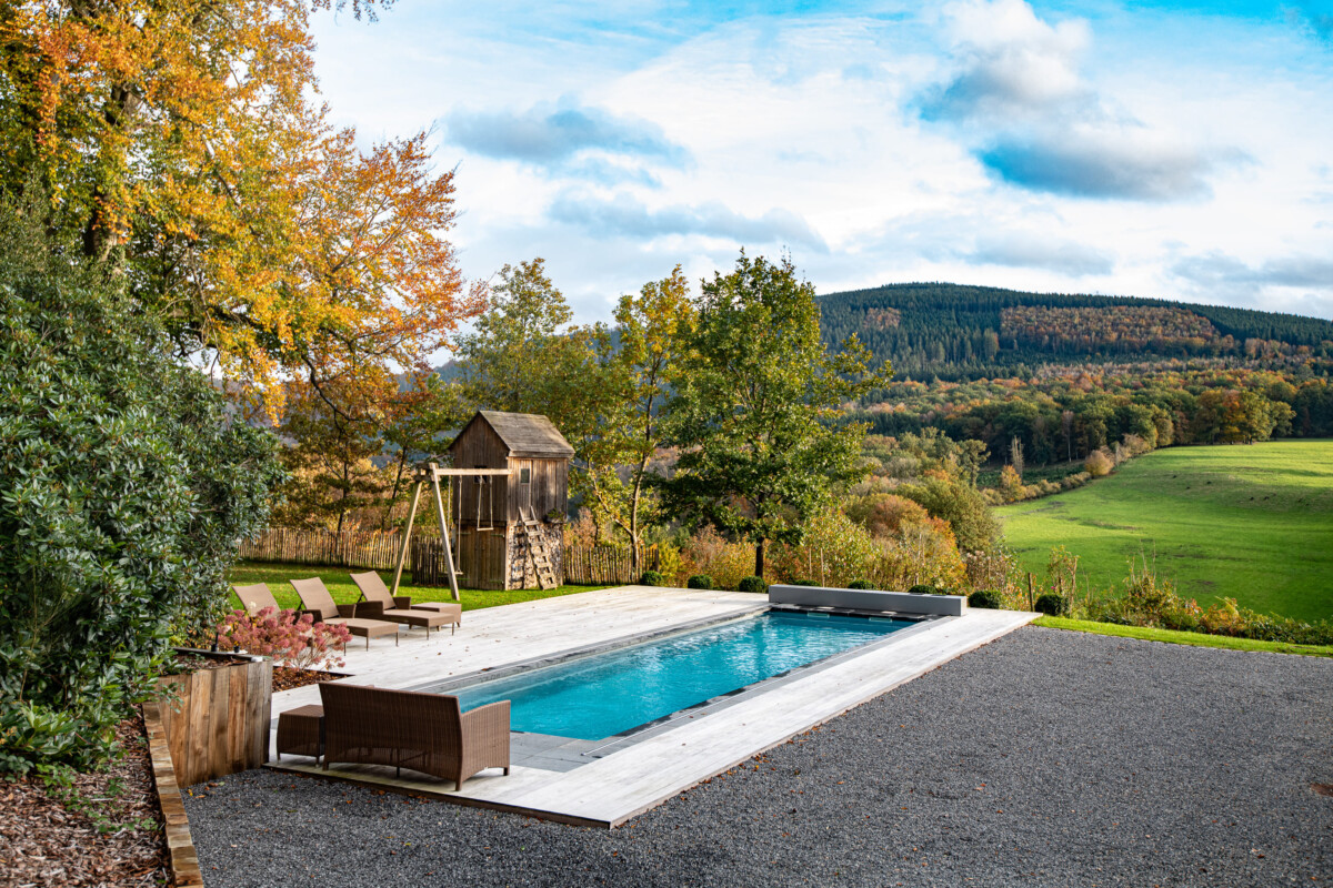 Une piscine rectangulaire entourée de chaises longues marron, avec un abri en bois et des arbres aux couleurs automnales en arrière-plan.