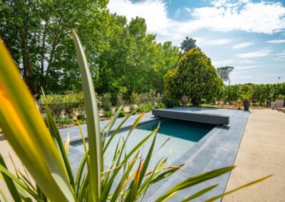 Piscine extérieure avec couverture grise entourée de végétation luxuriante et terrasse en pierre sous un ciel partiellement nuageux.