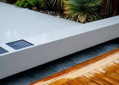Tropical_rain18-min Une couverture de piscine grise avec des panneaux solaires est entourée de plantes vertes et d'une terrasse en bois.