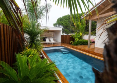 Tropical_rain27-min Piscine rectangulaire entourée de végétation tropicale, avec terrasse en bois et chaises blanches sous un abri ouvert.