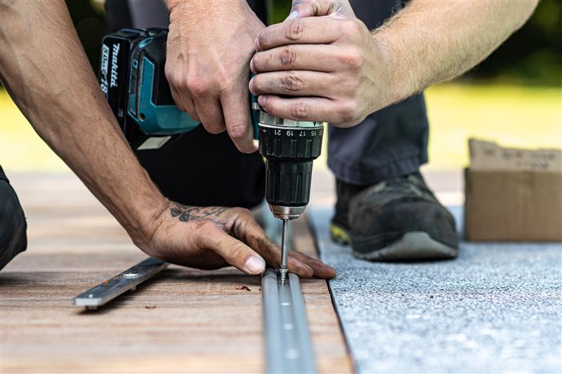Deux mains installent une vis avec une perceuse sur un rail métallique posé sur du bois, avec un carton à proximité.
