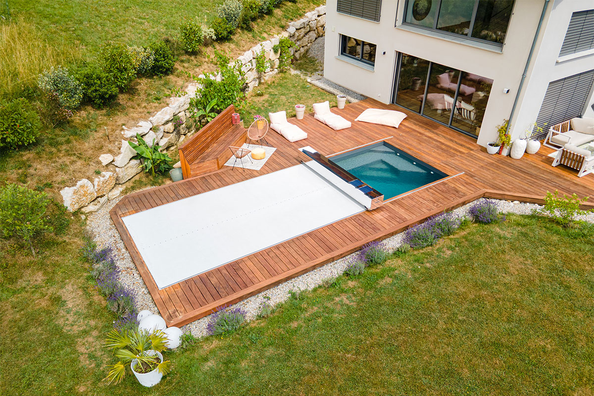 Piscine moderne avec terrasse en bois et couverture automatique fermée, intégrée dans un jardin paysager.