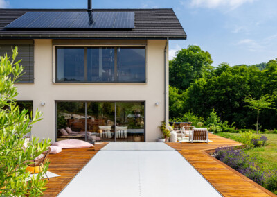 Maison contemporaine avec grandes baies vitrées et terrasse en bois équipée d’une piscine avec couverture fermée.