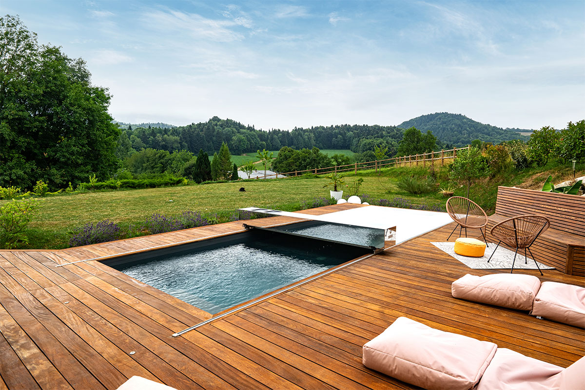Piscine moderne avec terrasse en bois et volet automatique partiellement ouvert, donnant sur un grand jardin verdoyant et vallonné.