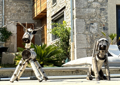 Détail de deux sculptures décoratives en bois et métal représentant des animaux, installées au bord d’une piscine devant une maison en pierre.
