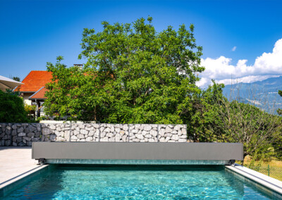 Piscine rectangulaire avec couverture Coverseal partiellement ouverte, devant un mur en pierre et un jardin verdoyant sous un ciel ensoleillé.