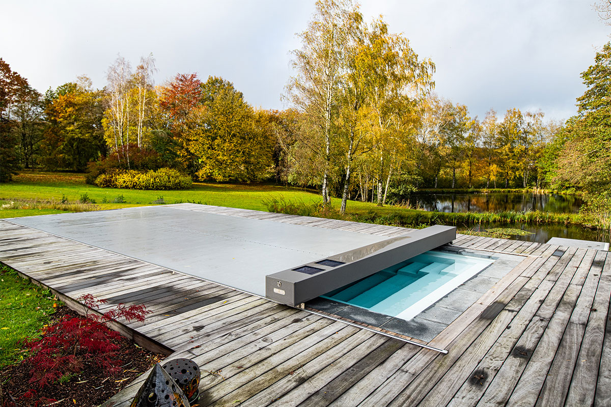 Piscine rectangulaire équipée d’une couverture Coverseal grise, entourée d’une terrasse en bois, avec un grand jardin verdoyant et un étang en arrière-plan.