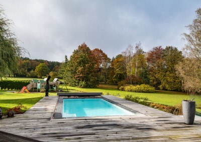 Pond7 Piscine turquoise entourée d’une terrasse en bois, avec aire de jeux pour enfants et jardin aux couleurs d’automne en arrière-plan.