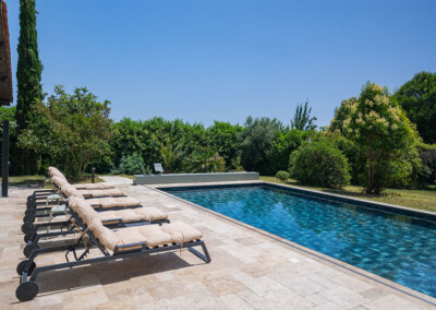 Perspective du bord opposé de la terrasse, montrant la piscine bleue entourée de transats et intégrée dans un environnement verdoyant.